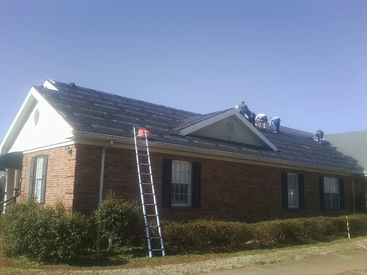 Crew tearing shingles off a brick ranch roof with a ladder set — Metal Roof Repair work by our crew in Jarratt